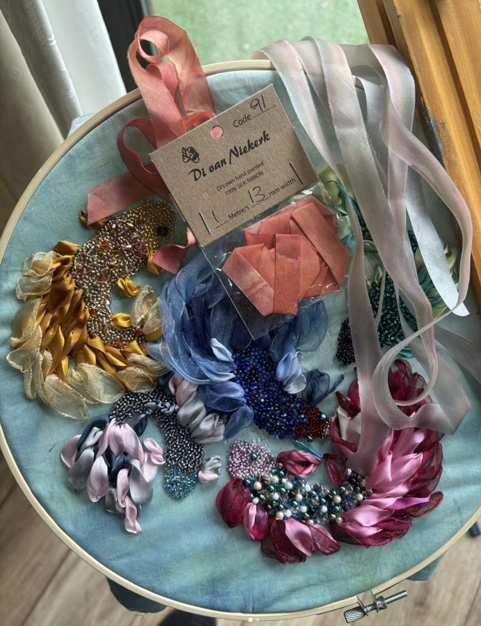 Lengths of hand-painted silk ribbon, beadwork and ribbon flowers laid out across a pale blue embroidery hoop