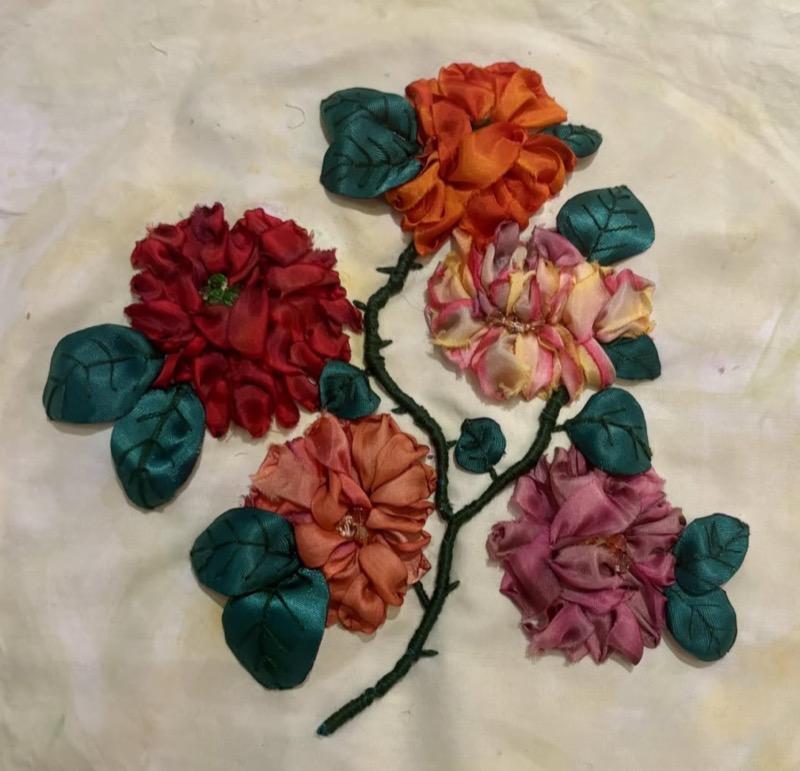 A spray of red, orange and pink silk ribbon roses with deep green leaves, on cream cloth